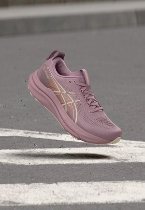 Lilla løbesko med beige detaljer ser ud til at springe over en grå asfaltvej med hvide markeringer.
