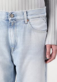 Jeans de denim azul claro con una textura desgastada, que cuentan con un solo bolsillo frontal y un cierre de botón metálico. La mano descansa en el costado.