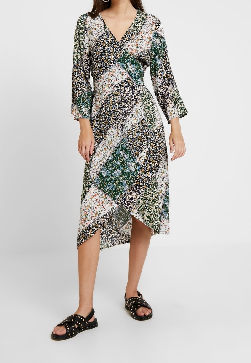 Vestido envolvente con estampado floral y mangas tres cuartos, en tonos de verde, blanco y navy; con dobladillo asimétrico; combinado con sandalias negras con tachuelas.
