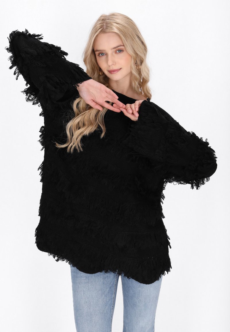 Zwarte oversized trui met gestructureerde franjes langs de mouwen en het lichaam, met een ronde halslijn. Gecombineerd met lichtblauwe jeans.