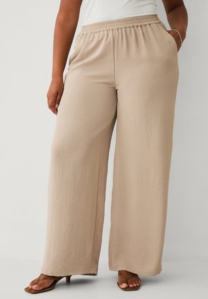 Kvinde iført beige bukser med vide ben, elastisk talje og lommer, kombineret med brune åbne hælede sandaler.