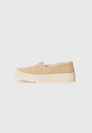 Beige kanvas slip-on sko med en teksturert overflate, rund tå og en tykk, kremfarget gummisåle. Ingen synlige snørebånd eller hardware.