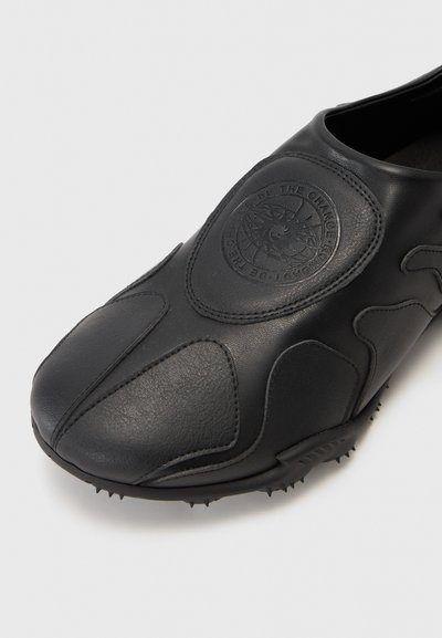 Zapato de cuero negro con superficie texturizada, diseño moldeado, parche redondo de logo en la parte superior y picos de goma en la suela para tracción.