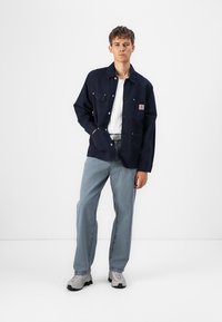 Chaqueta de botonadura azul marino con dos bolsillos en el pecho y parche de marca, combinada con jeans azul claros y zapatillas grises.