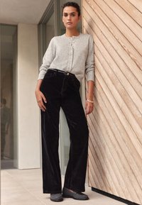 Cárdigan gris jaspeado con cierre de botones acompañado de pantalones anchos de pana negros y zapatos grises sin cordones sobre un fondo de madera.