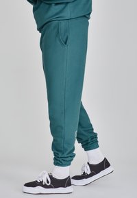 Teal sweatpants med sidfickor och elastiska muddar, i kombination med svarta sneakers och vita strumpor. Tyget verkar mjukt och slätt.