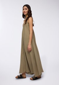 Vestito lungo senza maniche di colore verde oliva con una forma fluida, realizzato in tessuto testurizzato. Abbinato a sandali neri con dettagli metallici.