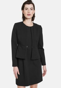 Schwarze taillierte Jacke mit rundem Ausschnitt, strukturierten Einsätzen und einem Schößchen, kombiniert mit einem schlichten schwarzen Kleid. Glatte Stoffoberfläche.