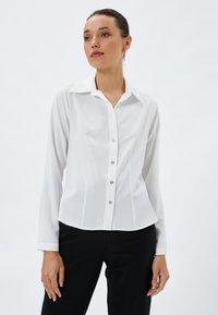 Hvid langærmet bluse med klassisk krave, frontlukning med sølvfarvede knapper og en skræddersyet pasform. Glat stoftekstur.