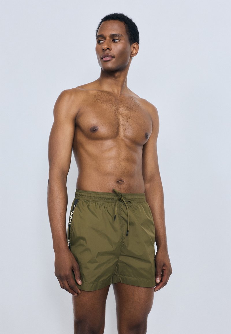 Oliivirohelised ujumispüksid kergest, läikivast materjalist, millel on elastne vöökoht ja pael. Tunnuseks on küljes triibud ja lõdvestunud lõige.