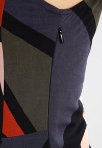 Närbild av geometriskt mönstrad tyg i marinblått, svart, olivgrönt och rött med en liten svart dragkedja på ett plagg.