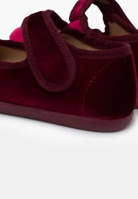 Pantuflas de terciopelo burdeos con punta redonda, que cuentan con una correa de Velcro y un interior suave. La suela es texturizada para mayor agarre.