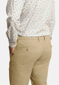 Pantalones caqui con una textura suave, con bolsillos traseros con detalles de botón, combinados con una camisa de color claro con estampado.
