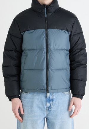 Homme portant une doudoune à blocs de couleurs noire et grise avec une fermeture éclair frontale, assortie à un jean bleu clair, debout devant un fond uni.