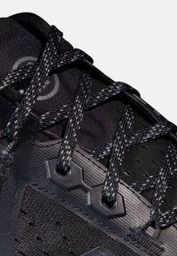 Svart sportskor med texturerad syntetisk ovandel, platta snörningar och hexagonala ögla-accenter. Har en ventilerande nätdesign.