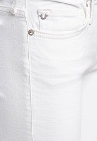 Close-up van een witte denim jeanszak met zichtbare metalen klinknagel en een klein metalen merkje aan de rand van de zak.