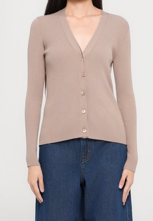 Femme portant un cardigan beige côtelé à manches longues boutonné et un jean bleu foncé à jambes larges, debout devant un fond uni.