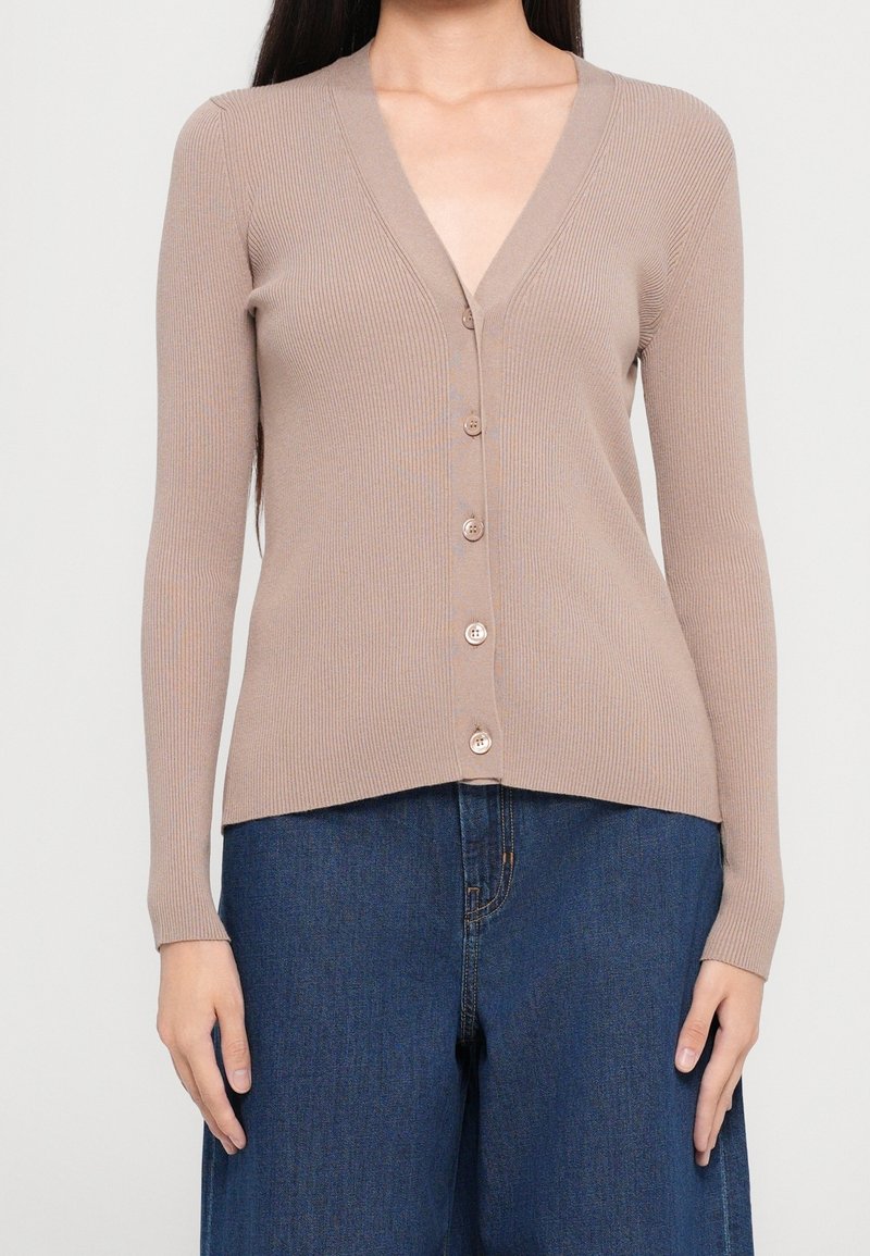 Femme portant un cardigan beige côtelé à manches longues boutonné et un jean bleu foncé à jambes larges, debout devant un fond uni.