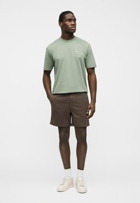 Homme debout les mains dans les poches d'un short marron, portant une chemise verte à manches courtes, des chaussettes blanches et des baskets beige sur un fond uni.