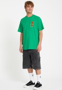 T-shirt en coton vert à manches courtes et avec un graphique sur le devant ; associé à un short en denim noir. Le modèle porte des baskets noires et des chaussettes blanches.