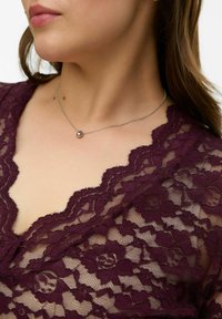 Bordeauxrode kanten top met een gebogen halslijn met bloemmotieven. Een dun zilveren kettinkje met een bolvormige hanger rust op het sleutelbeen.