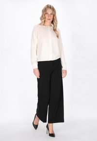 Crème kleurige cardigan met een gestructureerd bovenpatroon, ronde halslijn, knoopsluiting en lange mouwen, gecombineerd met hoge zwarte broeken.