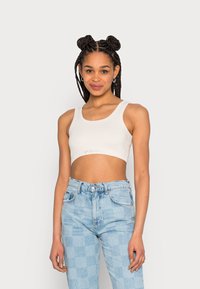 Top cropped bege claro com decote redondo e alças largas, combinado com jeans de cintura alta azul claro com padrão de xadrez. Design minimalista.