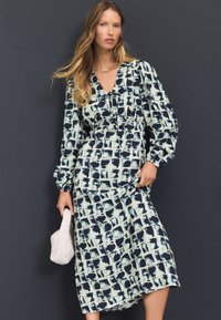 Robe à manches longues avec un motif à carreaux bleu marine et bleu clair, encolure en V, détails froncés et silhouette fluide. Tenant un sac blanc.