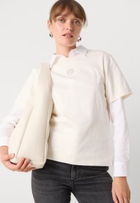 Maglietta corta crema con colletto a giro, caratterizzata da un logo ricamato. Abbinata a una camicia bianca con bottoni e jeans grigi.