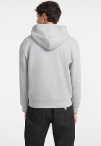 Sudadera gris con capucha y textura suave, con puños y dobladillo acanalados. Combinada con jeans de mezclilla oscuros. Etiqueta del logo visible cerca de la cintura.