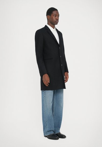 Un palton negru din lână cu un design cu un singur rând de nasturi, două butoane, buzunare laterale, purtat peste o pereche de blugi largi de culoare albastru deschis și pantofi negri.