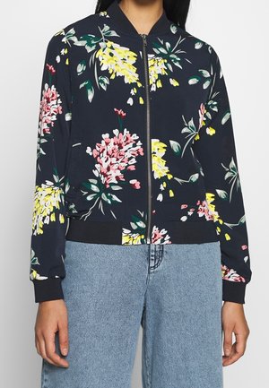 Femme portant une veste bomber bleu marine à imprimé floral rose et jaune, avec poignets et ourlet côtelés noirs, portée avec un jean en denim bleu clair.