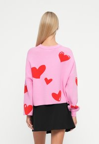 Pink knitted sweater with red heart patterns, long sleeves, and a crew neckline, worn over a black skirt. Smooth texture, cropped design.