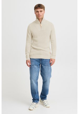 Beige knitted sweater with a high collar and ribbed texture, paired with blue denim jeans and light sneakers. Simple design, no visible patterns.