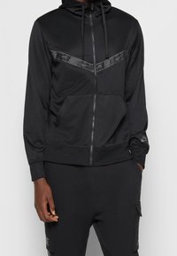 Svart zip hoodie i mjukt, lätt tyg. Har ett distinkt svart Nike-logoband över bröstet och en framficka.