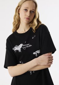Zwart katoenen t-shirt met witte grafische ontwerpen, waaronder tekst en veiligheidsspelden, met een losse pasvorm en korte mouwen.