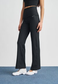 Μαύρο αθλητικό παντελόνι με ψηλή μέση, κατασκευασμένο από ελαστικό ύφασμα. Φέρει λογότυπο Nike σε λευκό στην περιοχή του γοφού. Συνοδεύεται από λευκά παπούτσια.