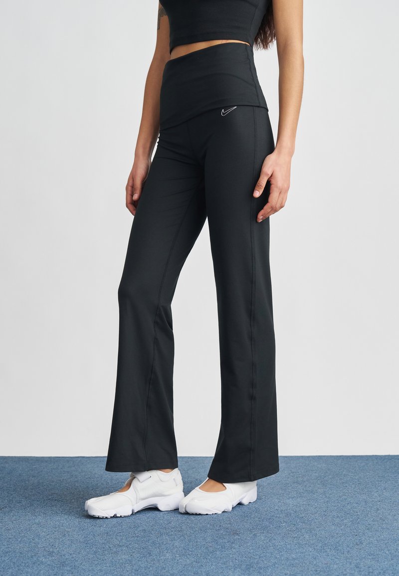 Μαύρο αθλητικό παντελόνι με ψηλή μέση, κατασκευασμένο από ελαστικό ύφασμα. Φέρει λογότυπο Nike σε λευκό στην περιοχή του γοφού. Συνοδεύεται από λευκά παπούτσια.