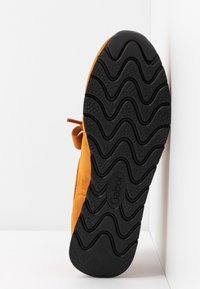 Chaussure jaune moutarde unique avec une semelle noire ornée de motifs ondulés et la marque "Gabor,", posée debout contre un mur blanc.