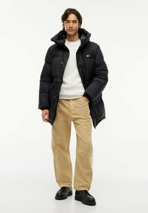 Junger Mann trägt eine schwarze Steppjacke, weißen Pullover, beige Hose und schwarze Schuhe und steht mit einer Hand in der Tasche vor weißem Hintergrund.