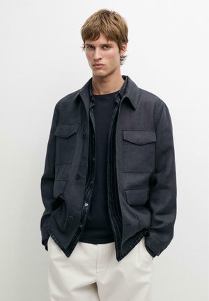 Ung mand med kort lyst brunt hår iført en sort jakke over en mørk skjorte og hvide bukser, stående foran en ensfarvet hvid baggrund.