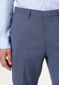 Maßgeschneiderte graue Hose mit einer dezenten Textur, flacher Vorderseite, schmaler Passform und Gürtelschlaufen. Kombiniert mit einem hellblauen Button-Up-Hemd.