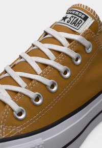 Κίτρινο καμβά παπούτσι με άσπρα κορδόνια, ασημένια μεταλλικά τρύπες και ετικέτα σε μαύρο και λευκό. Διαθέτει λευκή ελαστική σόλα με μαύρη ρίγα.