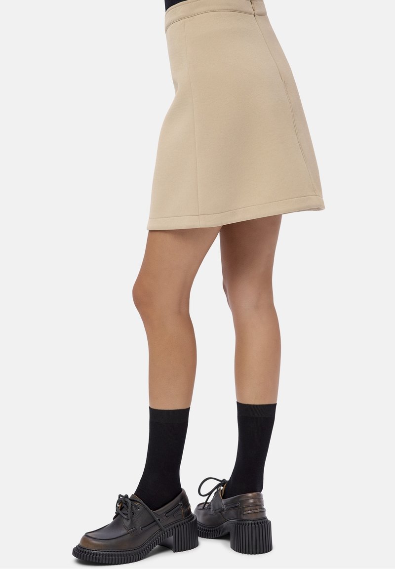 Mini-jupe beige en ligne A portée avec des chaussettes noires mi-mollet et des chaussures noires à lacets à talons épais, montrée de la mi-cuisse vers le bas.