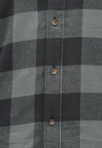 Camisa de flanela cinza e preta em padrão xadrez, com fecho de botões redondos castanhos na frente e tecido macio e texturizado.