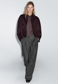 Bordeauxwollen vest met zwarte knopen, gedragen over een grijs overhemd, gecombineerd met grijze, gestreepte, wijde broekspijpen en zwarte schoenen.