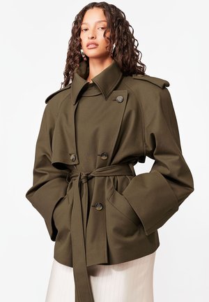 Kobieta o kręconych włosach, ubrana w oversize'owy oliwkowozielony płaszcz przeciwdeszczowy z szerokimi mankietami i wysokim kołnierzem, z rękami w kieszeniach, na białym tle.