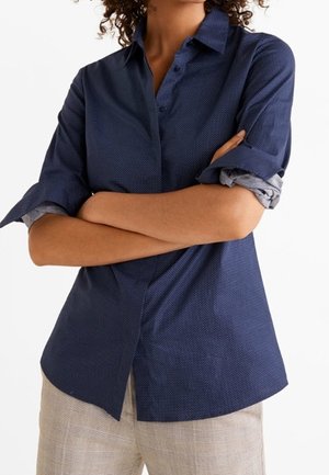 Person mit verschränkten Armen, die ein dunkelblaues langärmeliges Hemd mit hochgekrempelten Ärmeln und hellbeige Hose vor einem einfarbigen weißen Hintergrund trägt.