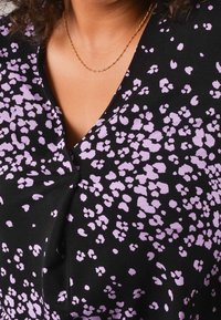 Černý textilní top s výstřihem do V, který má fialový květinový potisk. Textura vypadá jemně a design zahrnuje mírné nabírání kolem výstřihu.