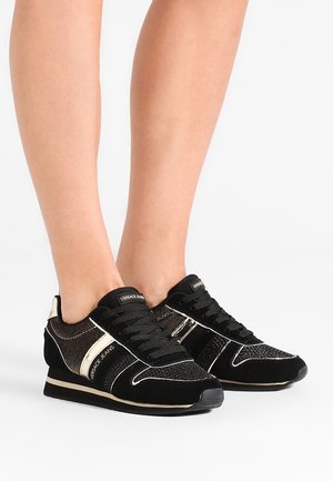 Baskets basses noires et dorées avec lacets, portées pieds nus, avec le nom de la marque "Versace Jeans" sur les côtés et la languette.